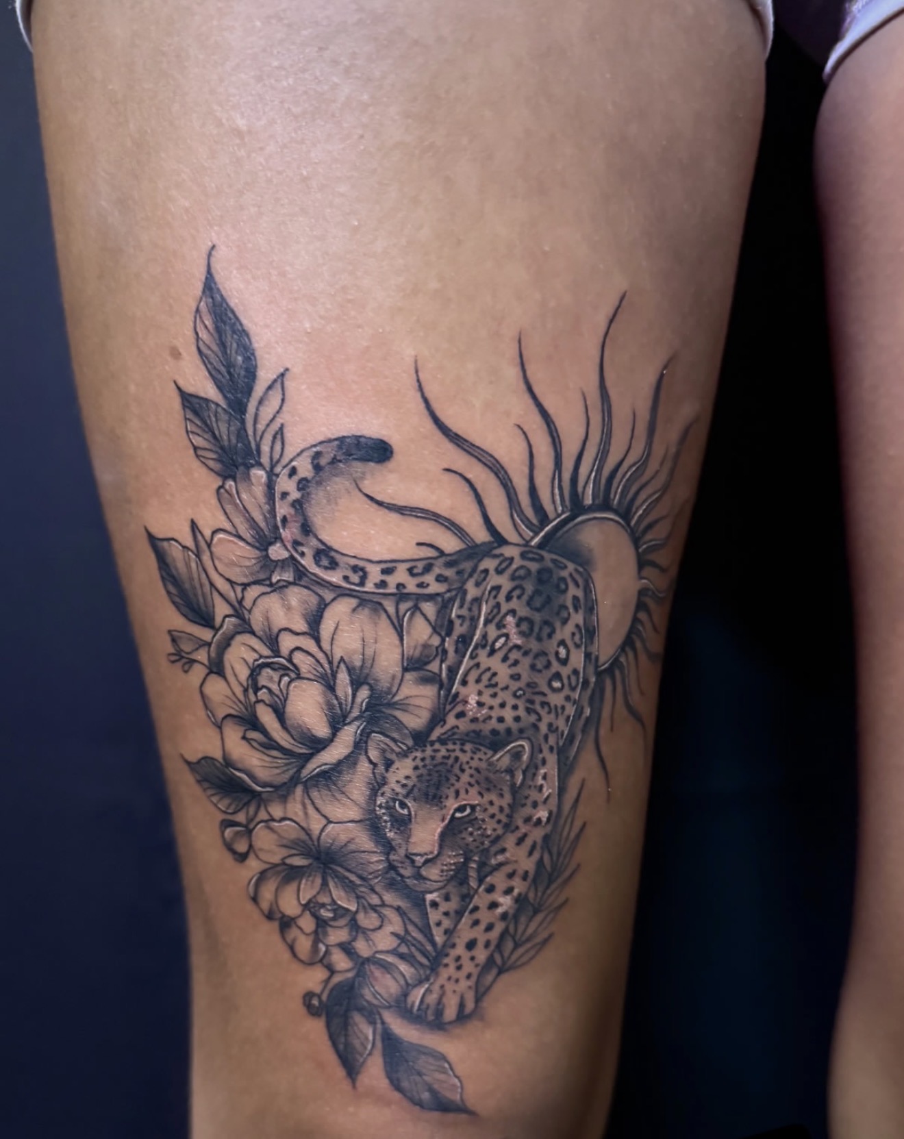 Black and gray thigh tattoo of a leopard among peonies with a radiating sun, by Peter Albert at Warpath Studio.