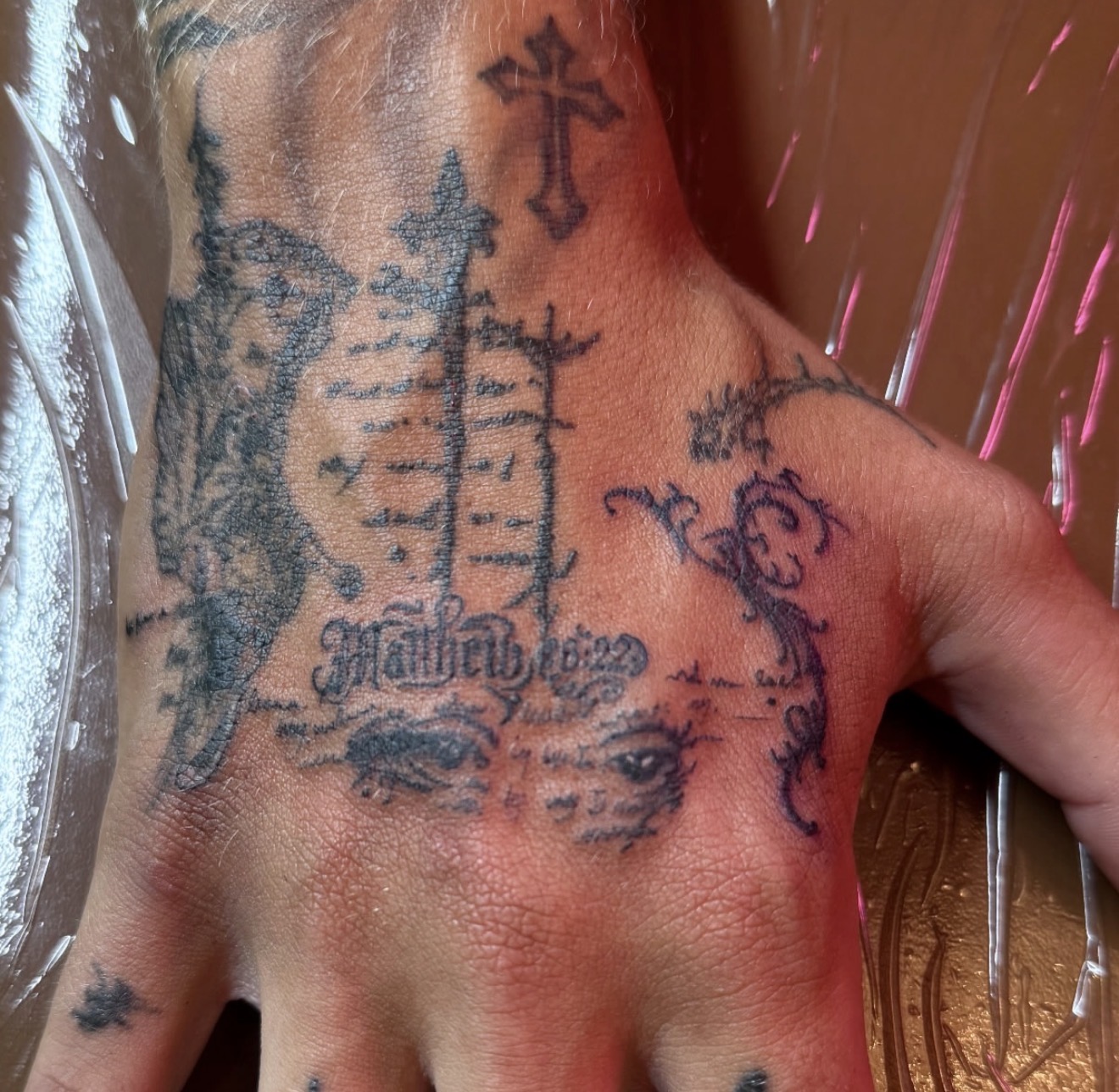 Freshly tattooed hand with crosses, ornamental linework and gothic scripture lettering, by Peter Albert at Warpath Studio.