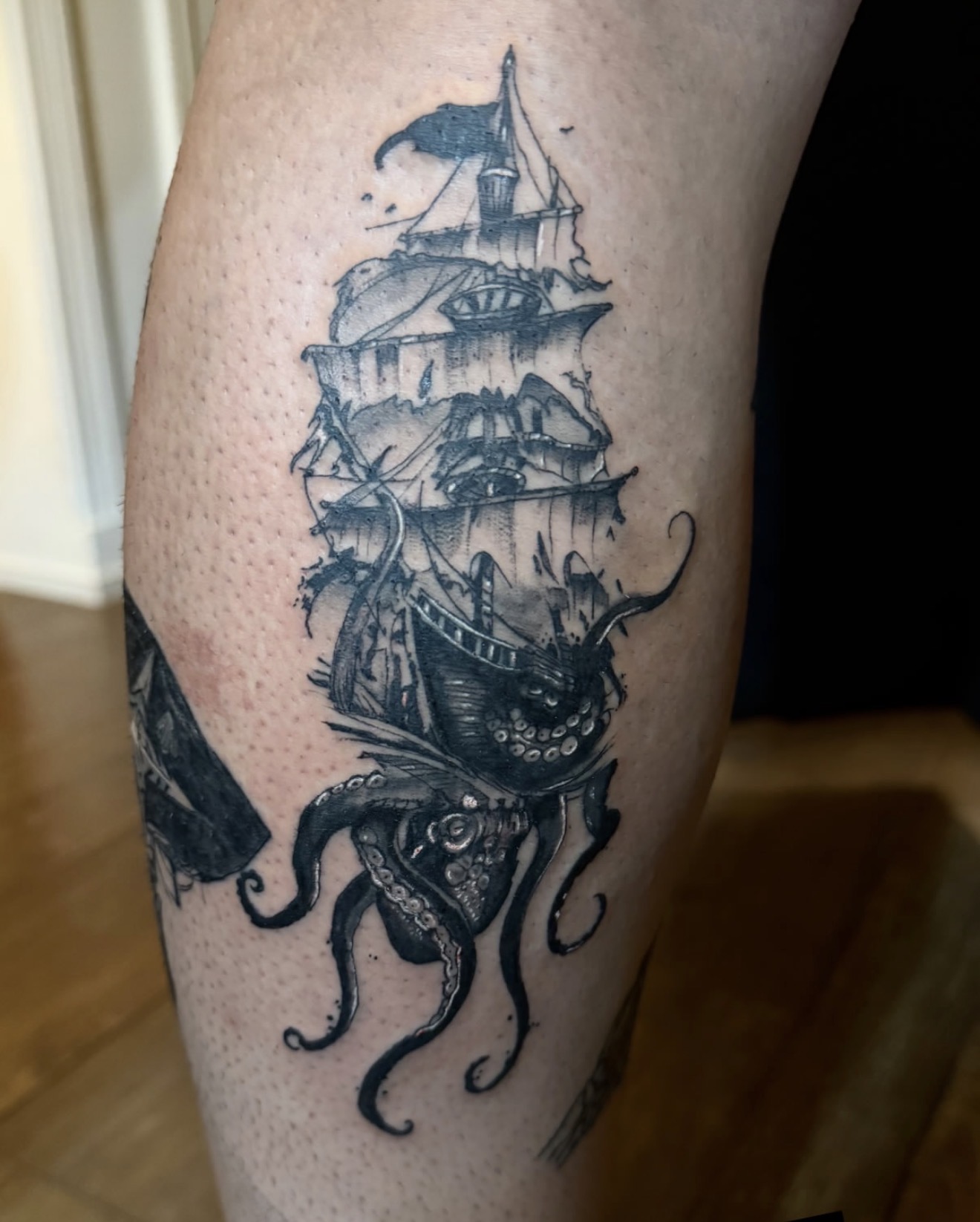 Black and gray calf tattoo of a tall ship being pulled under by a kraken, by Peter Albert at Warpath Studio.